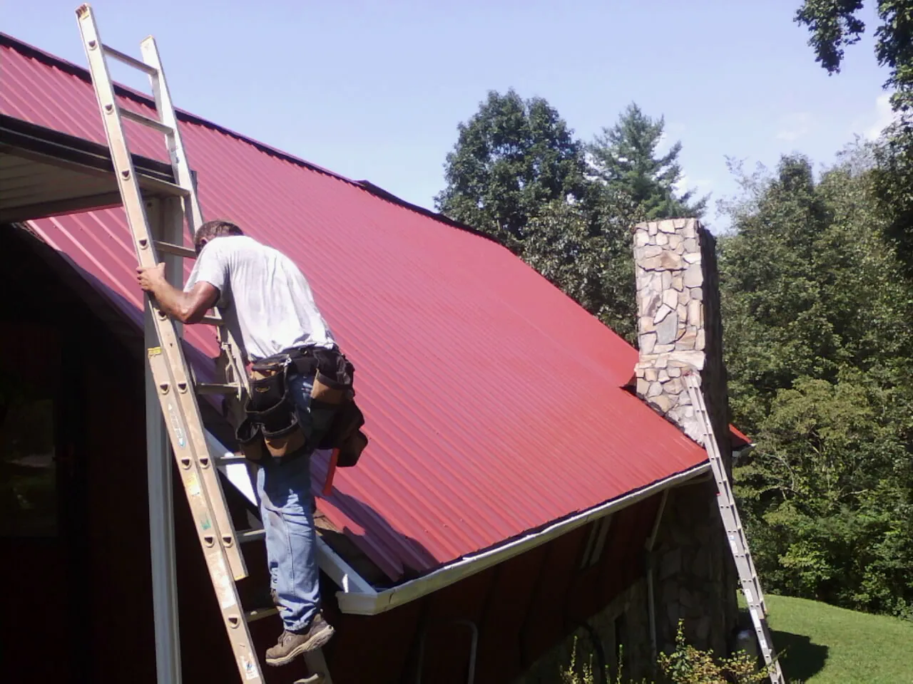 Roofer on a ladder beside a red metal roof and stone chimney — Soffit & Fascia Repair by Roofing Brothers in Amboy